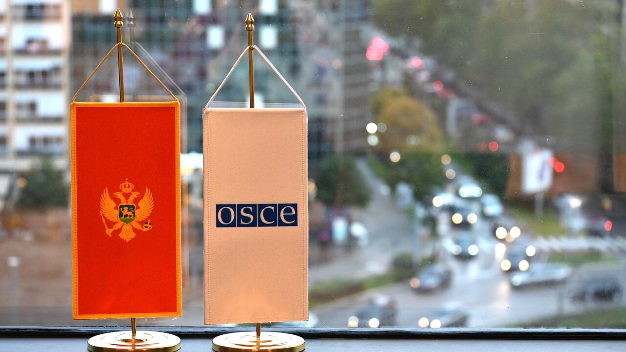 Two small table flags, one with Montenegro's emblem and the other with the OSCE logo, in front of a city street view.