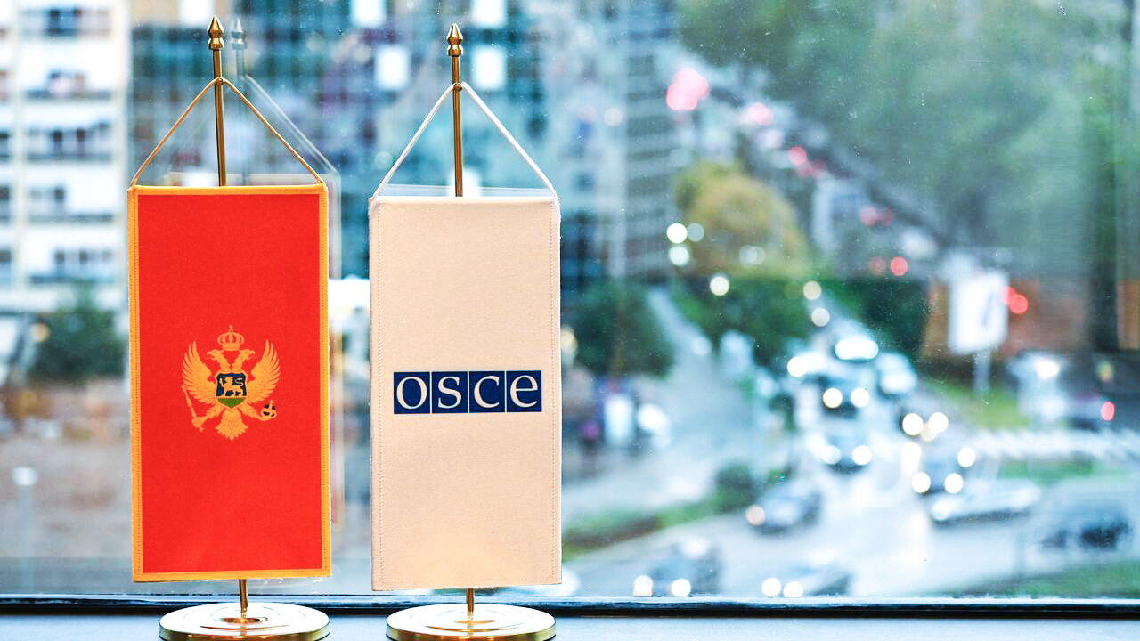 Two small table flags, one with Montenegro's emblem and the other with the OSCE logo, in front of a city street view.